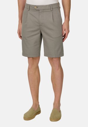 Mann trägt hellgraue knielange Shorts mit vorderen Falten und beigen Slippers vor einfarbigem Hintergrund.