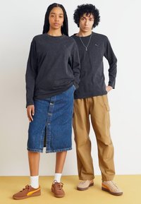 Tricouri negre cu mânecă lungă, fustă jeans tip creion cu fanta frontală, pantaloni cargo bej și pantofi sport maro cu accente albe, stând pe un fundal deschis.