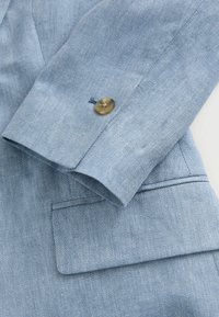 Svetle modrý džínový blazer s manžetou a zlatým knoflíkem. Obsahuje vzor herringbone, čisté švy a pečlivě přeložený klop.