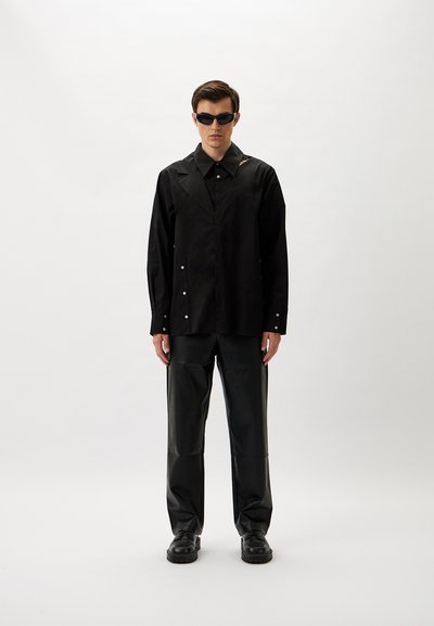 Schwarzes Langarmshirt mit weitem Kragen, Knopfdetails, kombiniert mit schwarzen, lederähnlichen Hosen und klobigen schwarzen Schuhen. Dunkle Sonnenbrille getragen.
