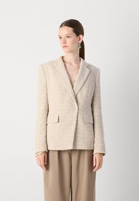 Beige rakenteellinen bleiseri suurella kauluksella ja etutaskuilla, yhdistettynä beigeen toppiin ja löysiin, vaaleanruskeisiin housuihin.