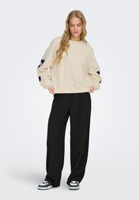 Femme aux cheveux blonds longs portant un sweat-shirt beige avec des imprimés noirs sur les manches, un pantalon noir ample et des baskets noir et blanc, debout.