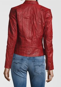 Frau in einer taillierten roten Lederjacke und blauen Jeans, von hinten vor einem schlichten hellen Hintergrund gezeigt.