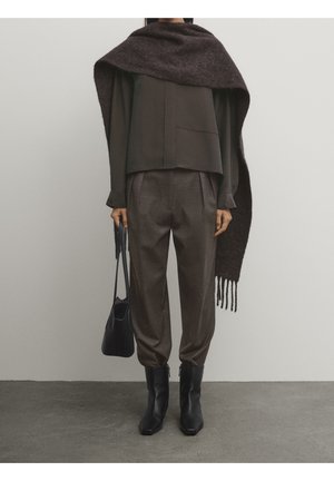 Person iført mørkebrune bukser, sorte støvler, en løs mørk top, stort mørkt halstørklæde draperet over skuldre, holder en sort håndtaske.