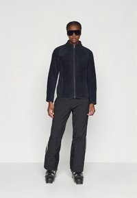 Granatowa kurtka polarowa z zamkiem błyskawicznym z przodu i wysokim kołnierzem, zestawiona z czarnymi spodniami z białymi paskami po bokach oraz czarnymi butami narciarskimi.