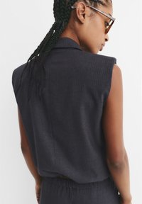 Ärmlös mörkgrå blazer med subtila randiga mönster, strukturerad axeldesign och slät tygtextur. Rückenansicht (bakvy) synlig.