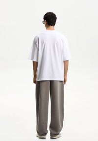 Witte oversized t-shirt met korte mouwen, gecombineerd met lichtgrijze wijde broek, van achteraf bekeken tegen een neutrale achtergrond.
