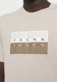 Μπεζ βαμβακερό T-shirt με γραφικό σχέδιο που φέρει το κείμενο "JACK & JONES" σε καφέ και λευκό, με υφασμάτινα ορθογώνια διακοσμητικά στοιχεία.