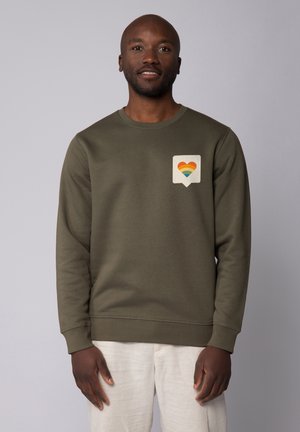 Olive green sweatshirt with crew neck, ribbed cuffs, and a patch featuring a rainbow heart inside a speech bubble. Soft texture, casual design.