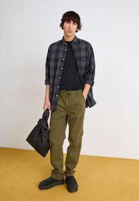 Black and gray plaid button-up shirt over a black t-shirt, olive green drawstring pants, black sturdy shoes, holding a dark puffy bag.
