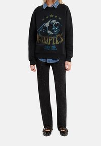 Person wearing black sweatshirt with graphic panther and stars, denim shirt underneath, black textured pants, and black pointed shoes with straps.