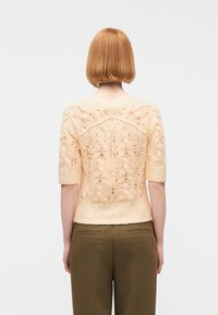 Άτομο με κοντά κόκκινα μαλλιά που φοράει ανοιχτό ροδακινί πλεκτό πουλόβερ κοντομάνικο και ελαιδί παντελόνι, στέκεται με την πλάτη γυρισμένη σε λευκό φόντο.