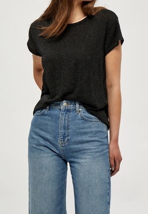 Kvinde iført en sort T-shirt med korte ærmer, stoppet ned i højtaljede blå jeans, stående med den ene hånd afslappet langs siden.
