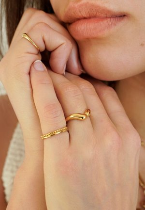 Close-up van de lippen en handen van een vrouw, waarbij drie gouden ringen met unieke ontwerpen op haar vingers te zien zijn.