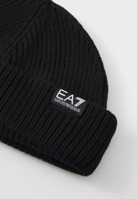 Černá pletená čepice s žebrováním a ohrnutým lemem, na které je připevněn obdélníkový černý štítek s bílým logem "EA7 EMPORIO ARMANI".