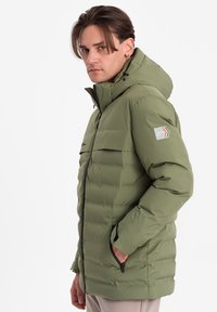 Olivově zelená nafouknutá bunda s kapucí, přední zip, vodorovný prošívaný design, boční kapsy a nášivka na levém rukávu.