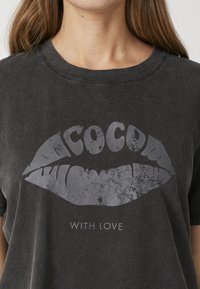 Juodas medvilnės marškinėliai su nusidėvėjusiu pilku lūpų grafiku ir užrašu „Cocoa“ bei „With Love“ po juo švariu šriftu.