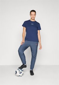 Mørk blå t-skjorte med trykt "PARIS" logo, grå joggebukser med rødt Nike-symbel, og svarte fotballsko, stående på en fotball.