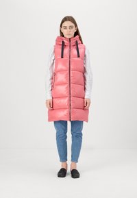 Gilet imbottito rosa con cappuccio, chiusura con zip frontale e coulisse nera. Presenta un motivo trapuntato e tasche laterali. Indossato sopra una maglietta a righe e jeans.