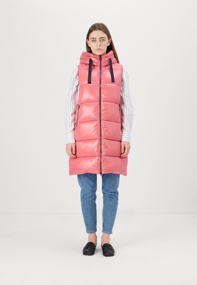 Gilet imbottito rosa con cappuccio, chiusura con zip frontale e coulisse nera. Presenta un motivo trapuntato e tasche laterali. Indossato sopra una maglietta a righe e jeans.