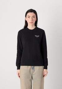 Kvinne med langt hår iført en svart hettegenser med logo, kombinert med lyse jeans, står mot en enkel hvit bakgrunn.