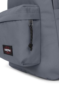 Mochila gris con un tejido duradero, que cuenta con un bolsillo frontal con cremallera y un tirador de cuerda negra. Incluye un parche de marca con acentos rojos.