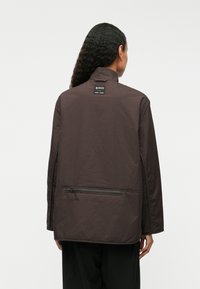 Temno rjav plašč z visokim ovratnikom, gladko teksturo, zadrgo na zadnjem žepu in oznako na vratu. Ohlapen kroj in zasnova.