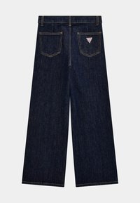 Weite dunkle Jeanshose mit vier Taschen, kontrastierender gelber Naht und einem kleinen dreieckigen Logo-Patch auf der Rückseite.