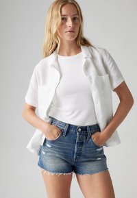 Balti trumpomis rankovėmis mygtukais užsegama palaidinė dėvima ant balto riboto tankinio viršaus, derinama su stipriai dėvėtomis džinsinėmis šortais su nusitrynusiomis kraštinėmis.