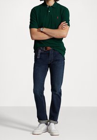 Ciemnozielona koszulka polo z czerwonym logo, niebieskie dżinsy i białe sneakersy. Pasek w paski. Model stoi z założonymi rękami.