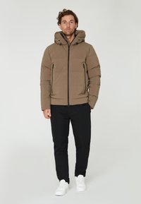 Beige vadderad jacka med huva och svarta dragkedjefickor, matchad med svarta byxor och vita sneakers på en enhetlig bakgrund.