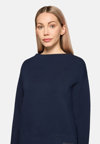 Sweatshirt bleu marine avec un col large, manches longues et une texture lisse. Présente un accent décoratif subtil près de l'ourlet.