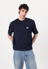 Navy cotton t-shirt with short sleeves and a crew neckline. Features a small rectangular patch with blue and white detailing on the chest.