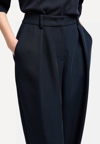 Pantalones de traje azul marino oscuro con un frente plisado, bolsillos laterales y una tela lisa y texturizada. Cintura ajustada con trabillas para cinturón.