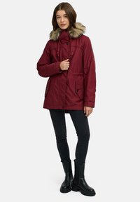 Parka bordeaux con cappuccio in pelliccia sintetica, chiusura anteriore a bottoni e coulisse, due tasche laterali, abbinato a jeans skinny neri e stivaletti.