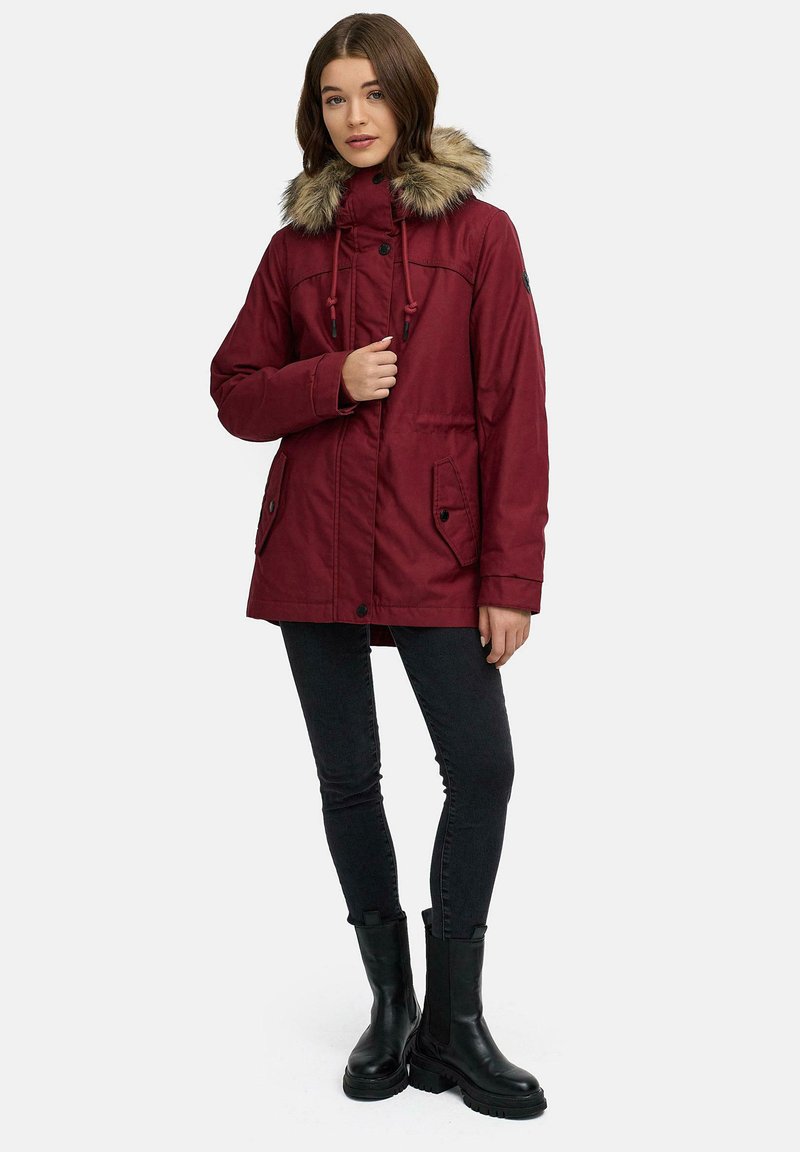 Parka bordeaux con cappuccio in pelliccia sintetica, chiusura anteriore a bottoni e coulisse, due tasche laterali, abbinato a jeans skinny neri e stivaletti.