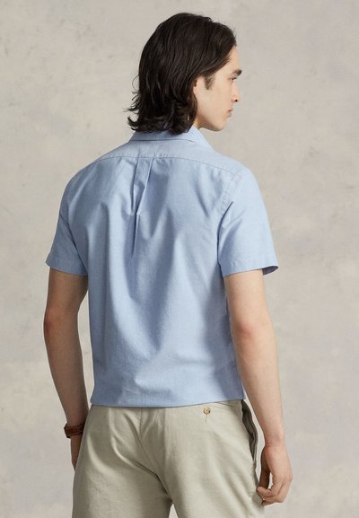 Camisa de manga curta azul-claro feita de algodão, com colarinho clássico e prega central nas costas. Combinada com calças bege claro.