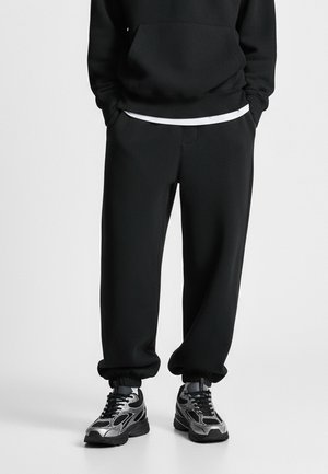 Schwarzer übergroßer Hoodie mit einer Kängurutasche, kombiniert mit passenden schwarzen tapered Sweatpants und eleganten silbernen Sneakers mit strukturiertem Finish.