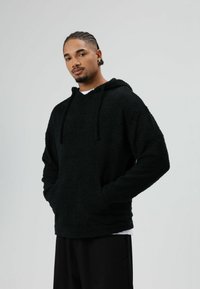 Schwarzer Fleece-Kapuzenpullover mit strukturierter Oberfläche, Fronttaschen und verstellbaren Kordeln. Über einem weißen Hemd getragen und mit schwarzen Hosen kombiniert.