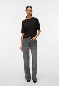 Vrouw staand in grijze, rechte, hoogtallige spijkerbroek, zwart kort mouw gebreid topje, bruine puntige schoenen, hand in de zak tegen witte achtergrond.