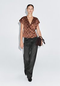 Bronze ternet mønstrede indpakningsbluse parret med sorte brede jeans. Modellen holder en mørkebrun ruskinds taske. Glat tekstur og tætsiddende design.