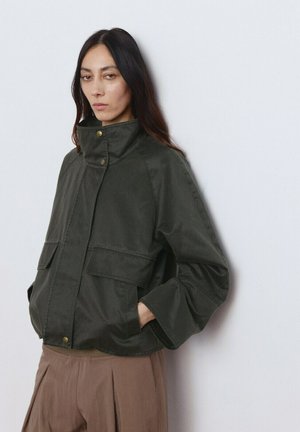 Mujer de cabello largo y oscuro con una chaqueta verde oscuro de cuello alto y bolsillos con solapa, y pantalones marrones plisados, de pie frente a una pared lisa.