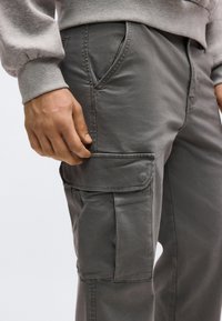 Άνδρας που φορά γκρι παντελόνι cargo με πλαϊνή τσέπη με καπάκι και γκρι φούτερ, στέκεται με το χέρι κοντά στην τσέπη.