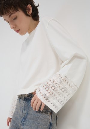 Top bianco a maniche lunghe realizzato in cotone con dettagli in pizzo alle maniche. I polsini presentano motivi floreali intricati e un design dal taglio rilassato.