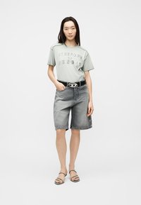 Γυναίκα με μακριά σκουρόχρωμα μαλλιά που φοράει ένα ανοιχτοπράσινο t-shirt "PROPERTY OF DIESEL", γκρι τζιν βερμούδα, μαύρη ζώνη και μαύρα σανδάλια.