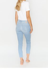 Žena s dlouhými blond vlasy, oblečená v krátkých modro-bílých pruhovaných džínách a bílé košili s krátkým rukávem, stojí naboso.