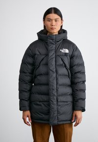 Giovane con capelli neri lisci che indossa un giubbotto imbottito nero con cappuccio e il logo bianco di The North Face, abbinato a pantaloni marroni.