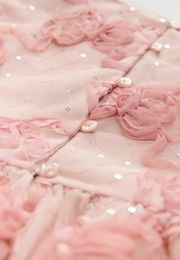 Tela de chifón rosa adornada con apliques florales en relieve, lentejuelas esparcidas y detalles de botones. Textura suave con un brillo sutil.