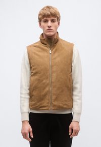 Rjava antilop jopič z visokim ovratnikom in zadrgo spredaj. Ima stranske žepe in je nošen preko svetlo sive rebraste majice.