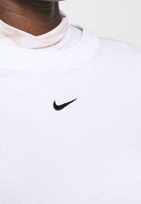 Vit bomullströja med rund halsringning och korta ärmar, prydd med en liten svart broderad Nike-logotyp på bröstet.